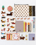 A flat-lay collection of dachshund-themed stationery and accessories on a white background, including a patterned pouch, sticker sheet, notebook, pens, keychain, and small decorative items.