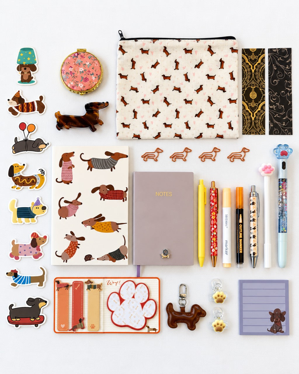 A flat-lay collection of dachshund-themed stationery and accessories on a white background, including a patterned pouch, sticker sheet, notebook, pens, keychain, and small decorative items.