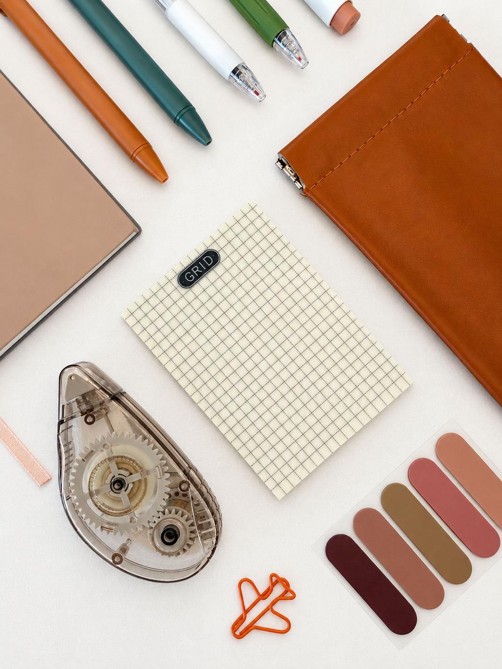 A styled flat-lay of stationery items on a white background, including a brown leather zip pouch, a beige grid notepad with a small black label reading 'GABBI', a clear tape dispenser, colored pens, a notebook, and a set of color swatches.