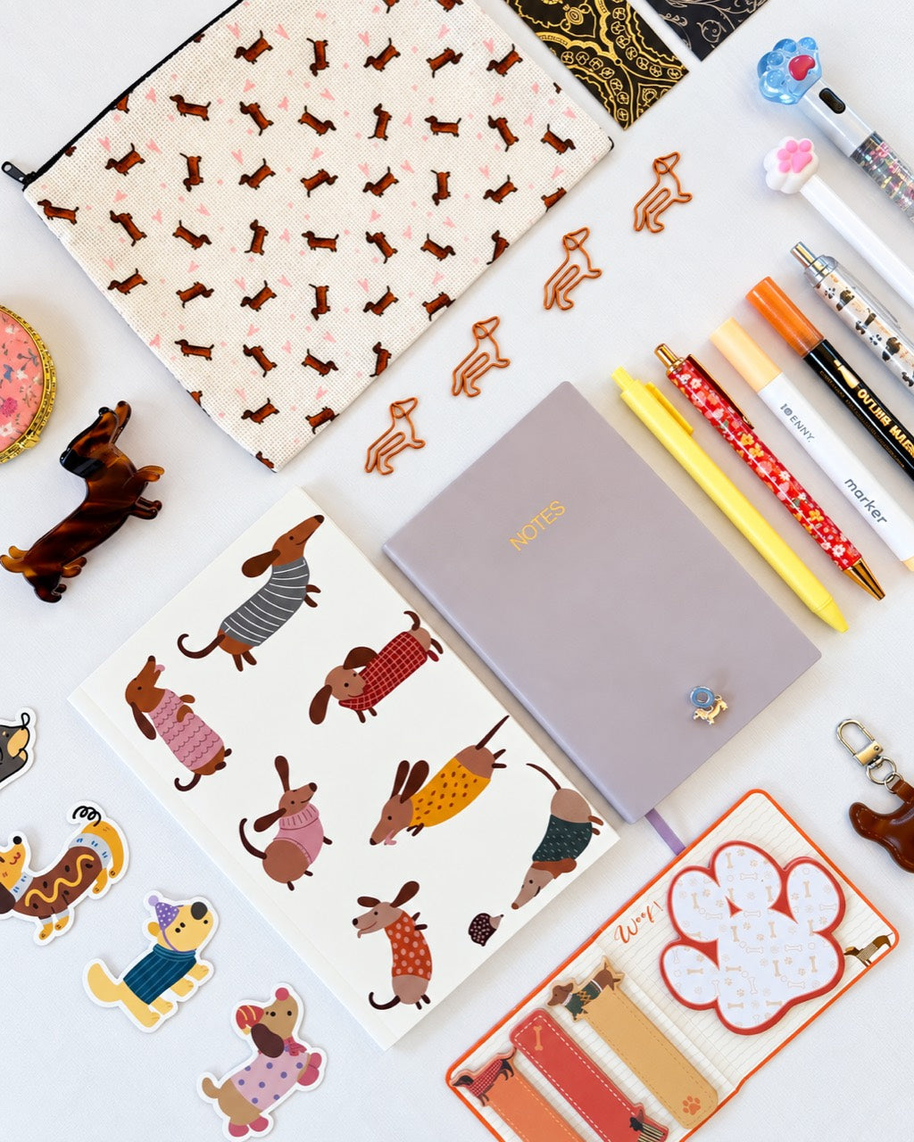 A styled flat-lay of stationery and accessories featuring dachshund illustrations. The set includes a lavender notebook with gold 'notes' lettering, a cream zippered pouch with brown dachshunds, a sticker sheet with colorful dachshund characters, yellow and orange pens, and a small leather wallet.