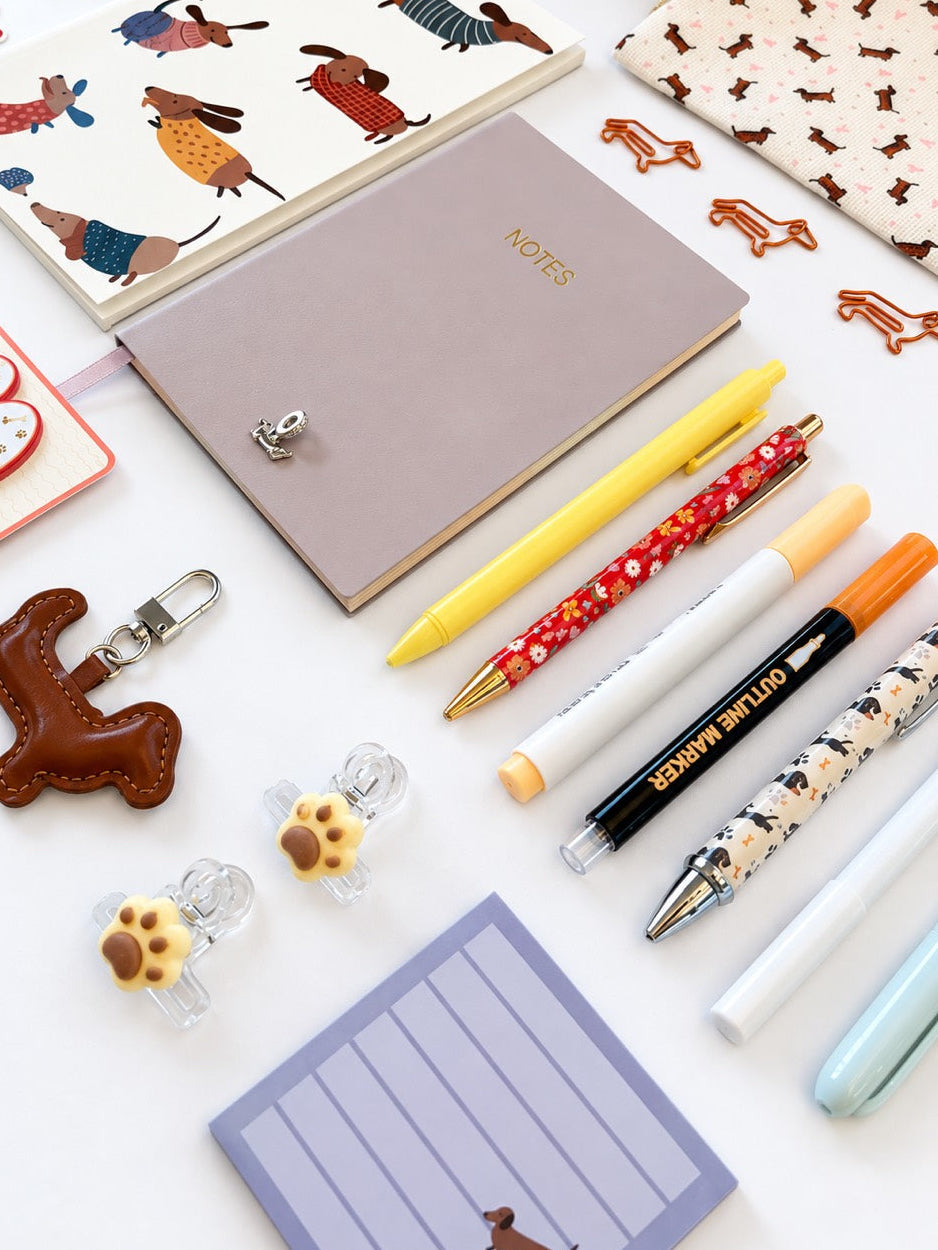 A styled flat-lay of dog-themed stationery and accessories on a white background, including patterned pens, a notebook, a notepad, a keychain, and small paw-shaped clips.